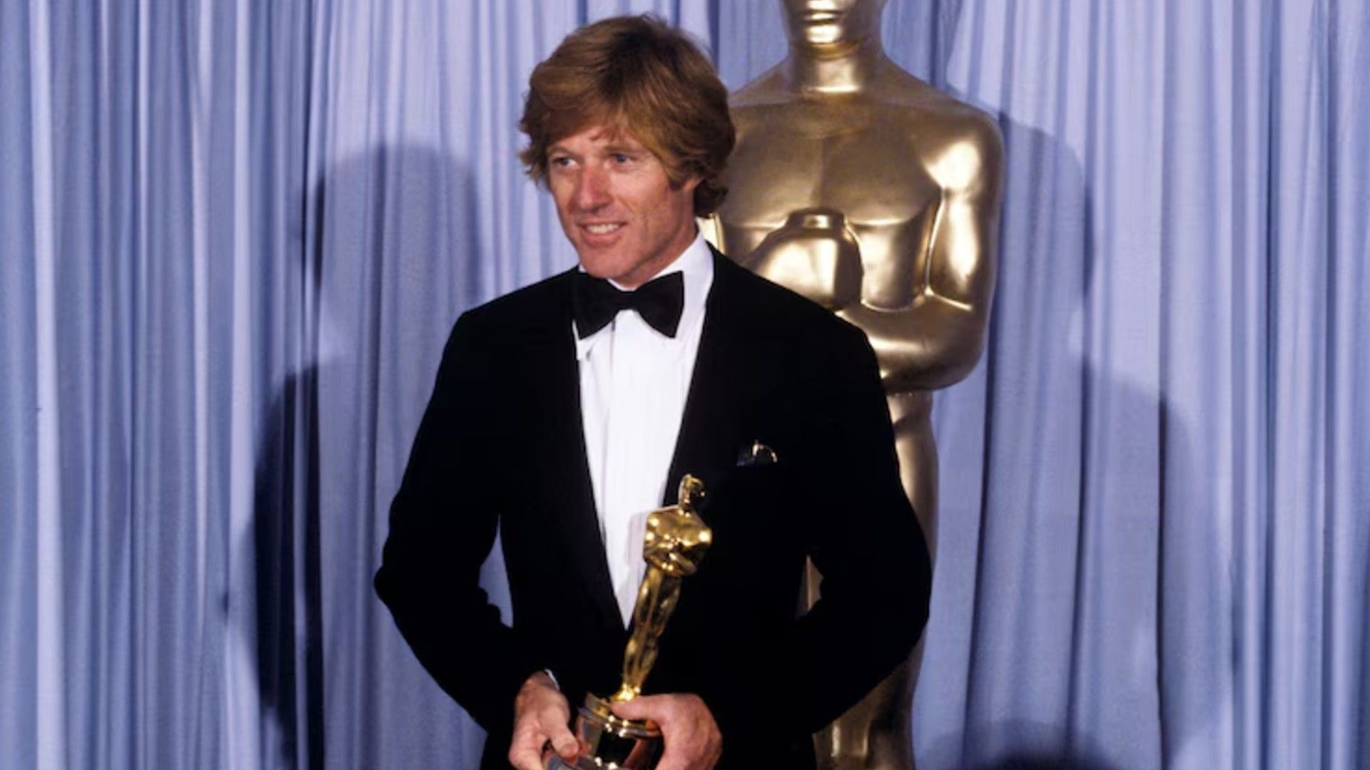 Robert Redford w garniturze stoi na scenie trzymając Oscara