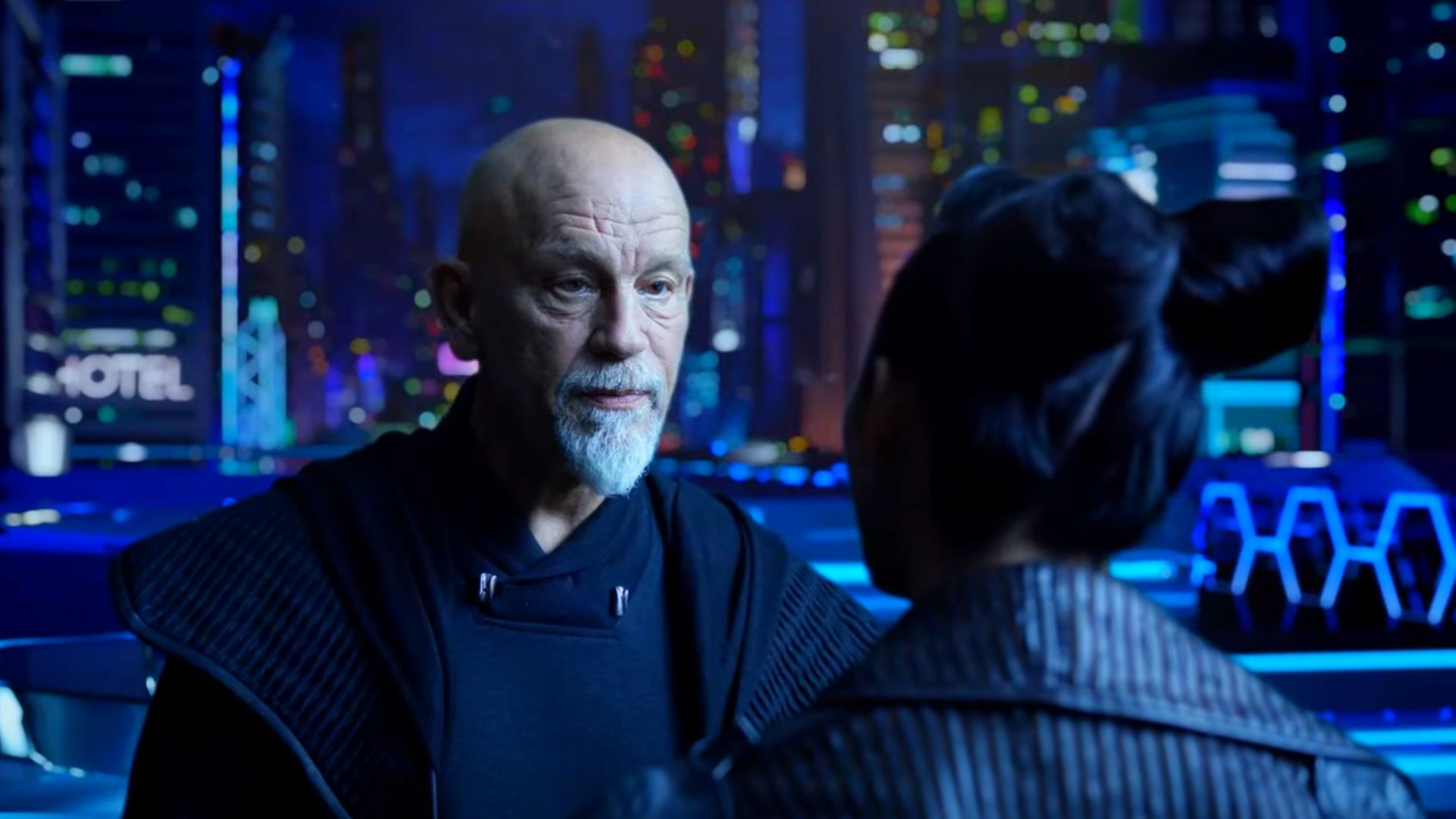 Aktor John Malkovich w futurystycznym stroju na tle futurystycznego nocnego miasta