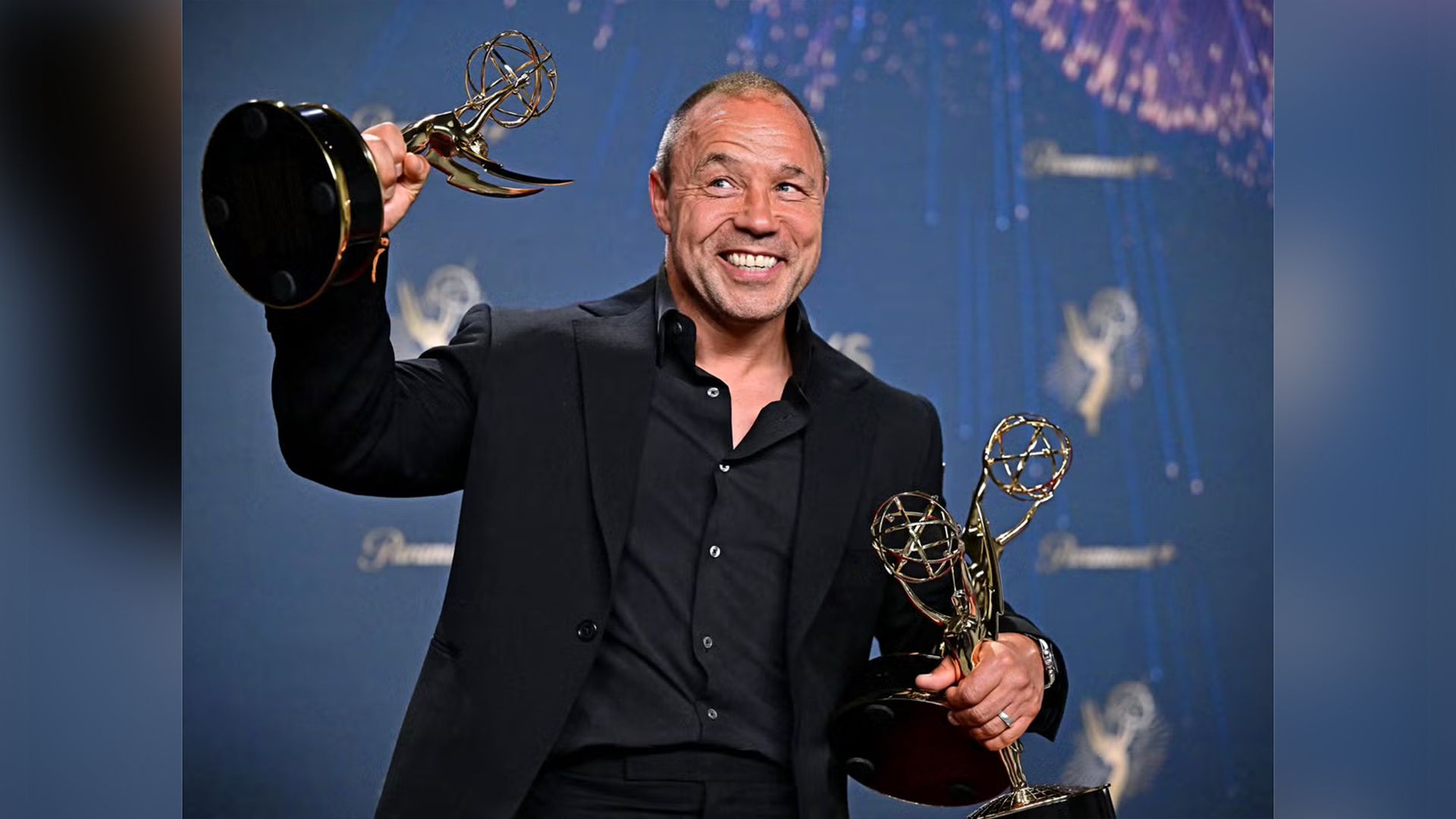 Stephen Graham w czarnym garniturze trzyma w rękach trzy złote statuetki Emmy, szeroko się uśmiechając