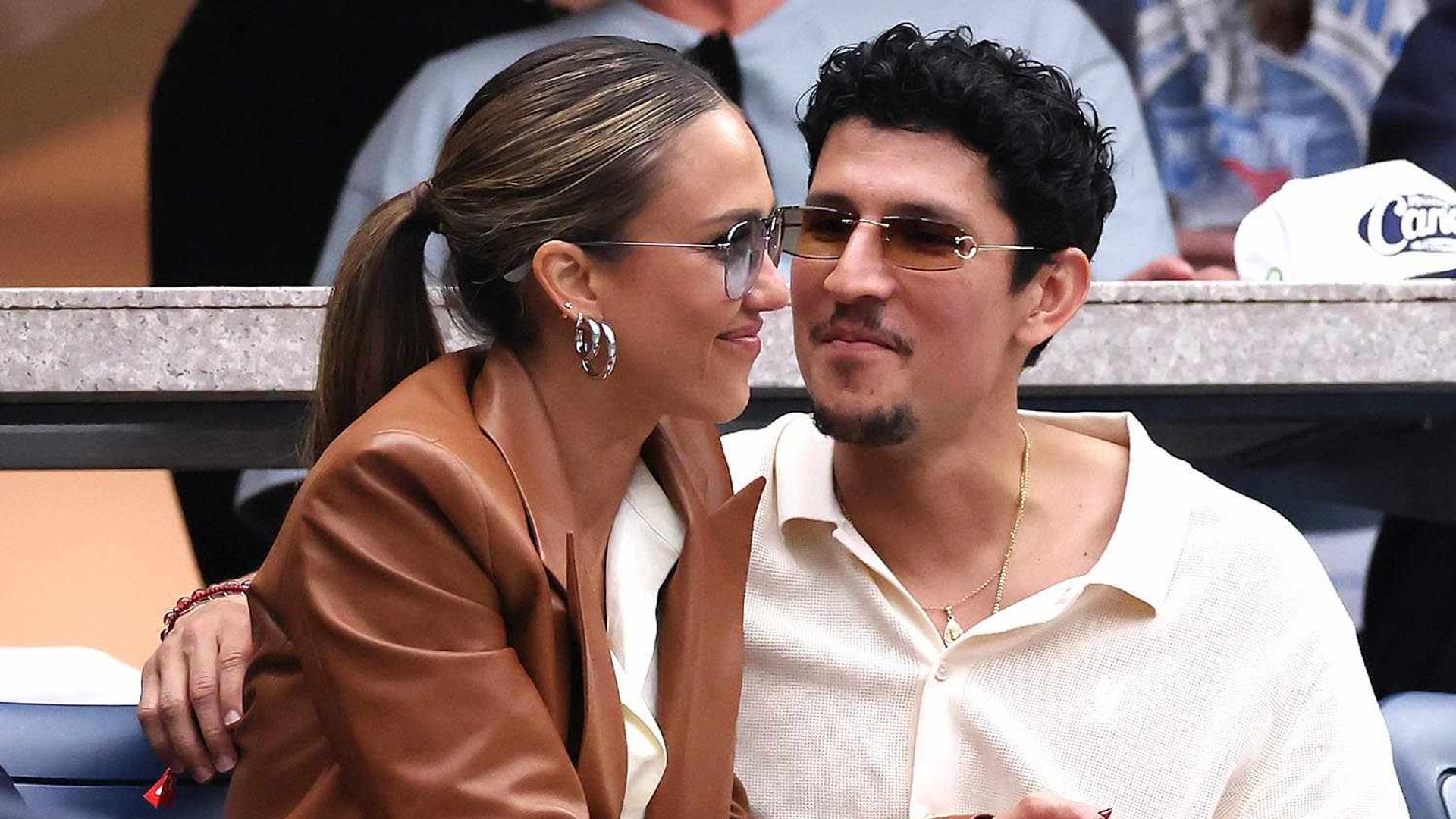 Jessica Alba i Danny Ramirez siedzący na trybunach podczas imprezy sportowej, obejmujący się nawzajem