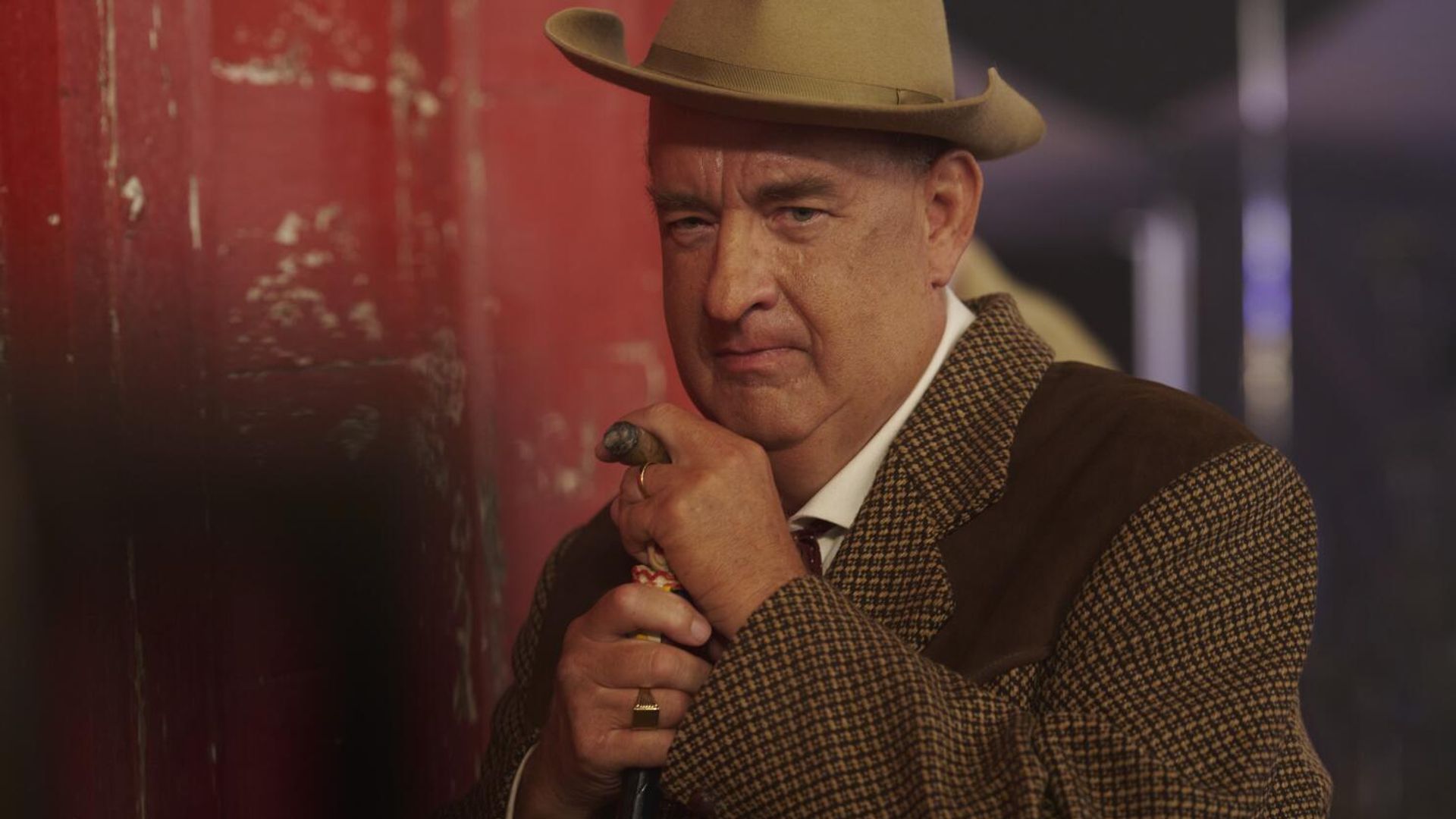Tom Hanks w brązowym garniturze i kapeluszu opiera się na lasce, patrząc poważnie w kamerę
