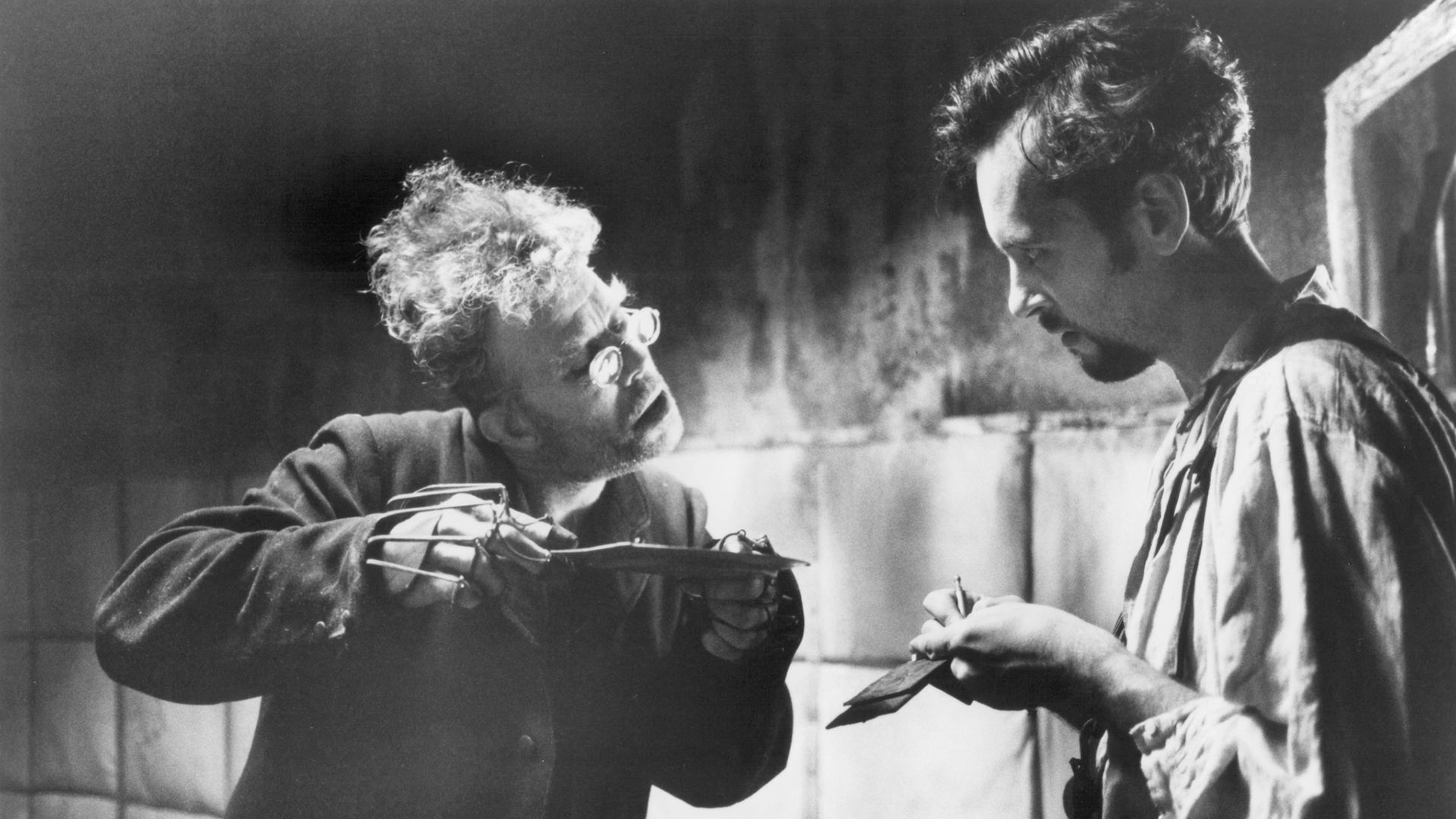 Tom Waits w okrągłych okularach i z szelkami na palcach trzyma spodek w dłoniach, obok niego stoi młody mężczyzna z notatnikiem