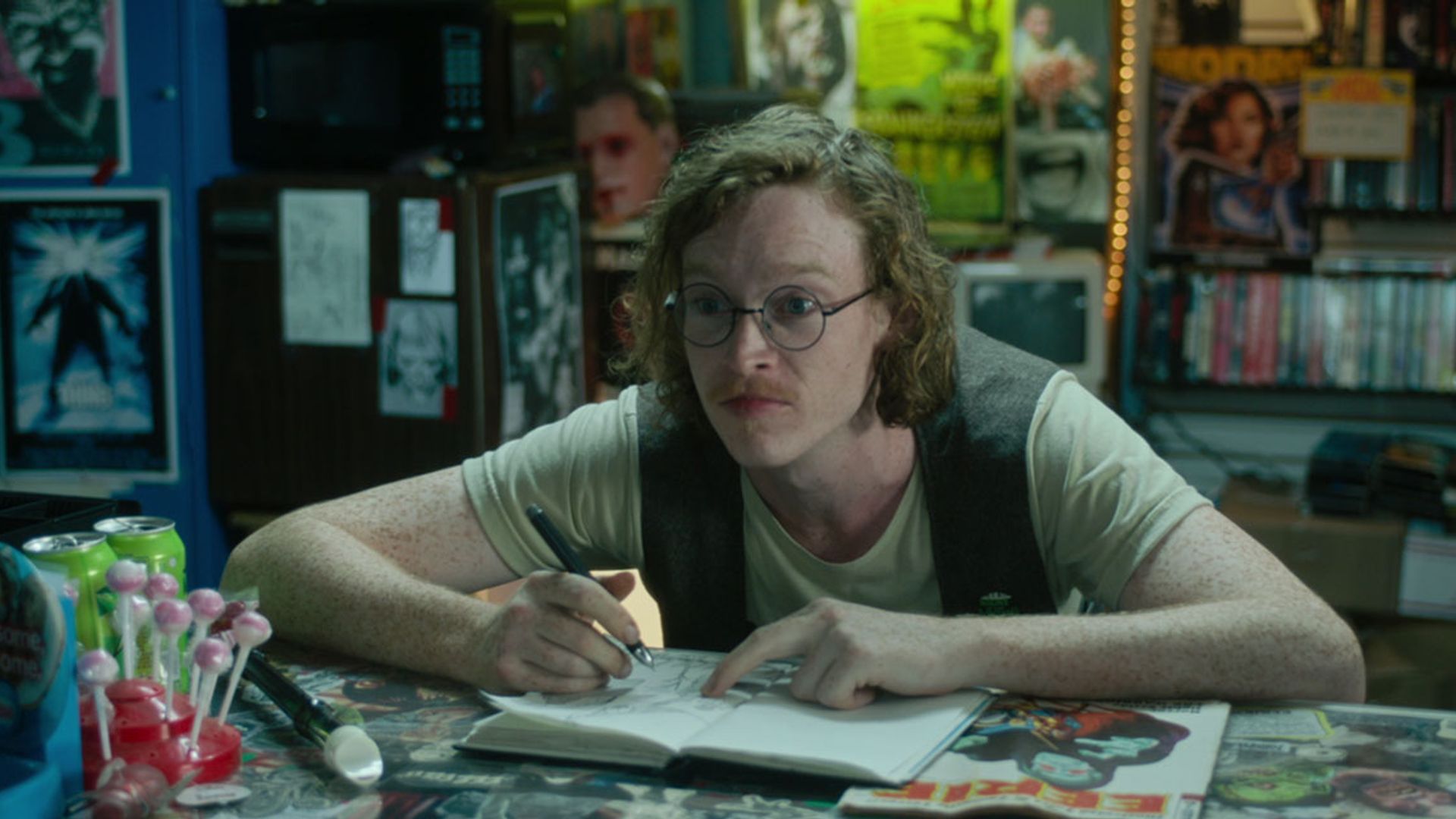 Caleb Landry Jones w okularach i kamizelce siedzi przy stole z notatnikiem, rysuje i patrzy w bok