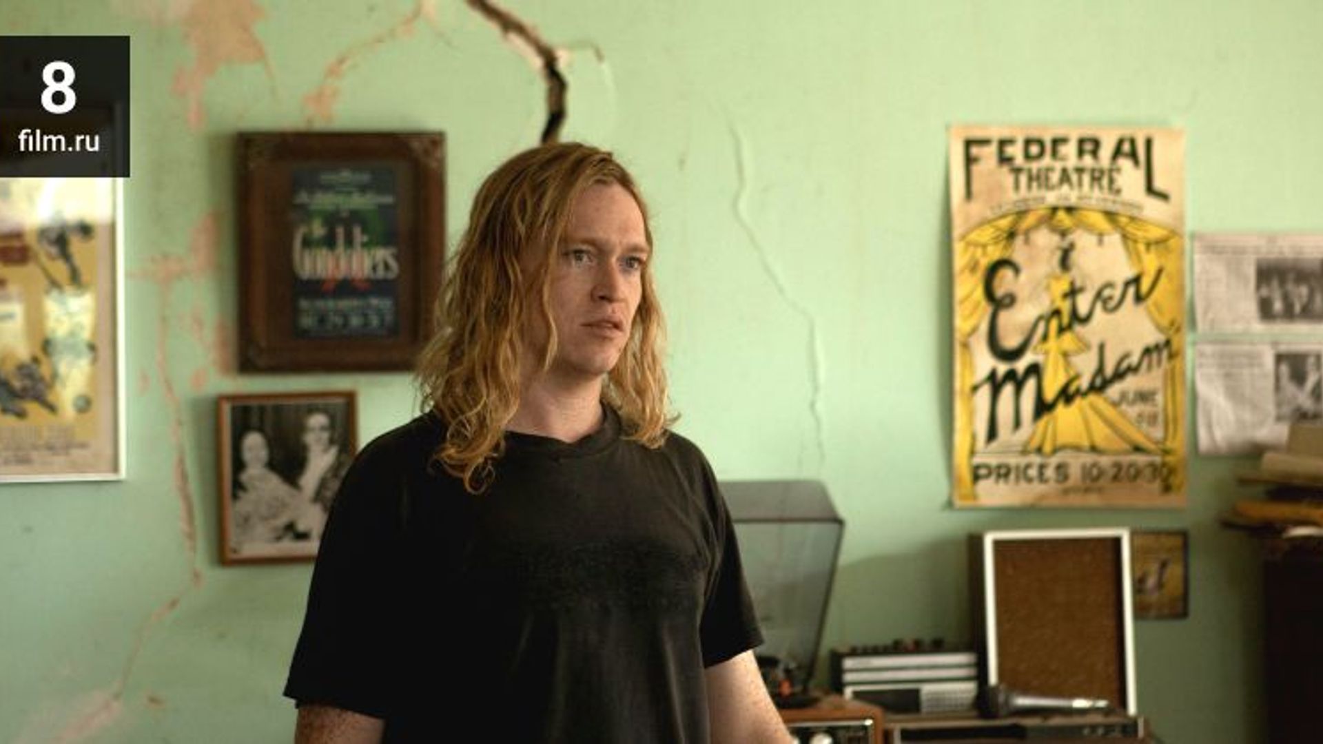 Caleb Landry Jones z długimi blond włosami w czarnej koszulce stoi w pokoju z plakatami na ścianie