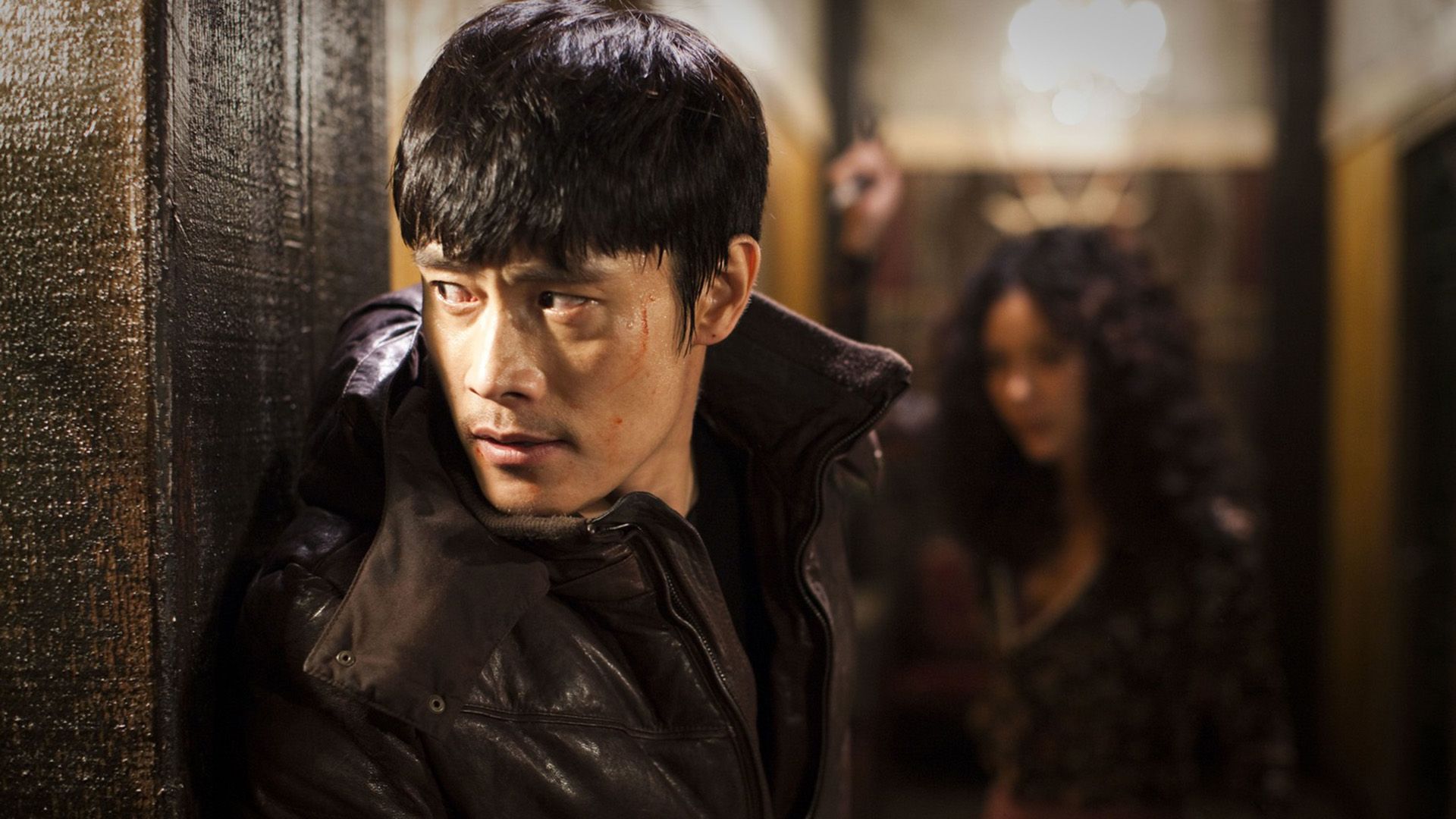 Lee Byung-hun w czarnej kurtce wygląda zza rogu budynku