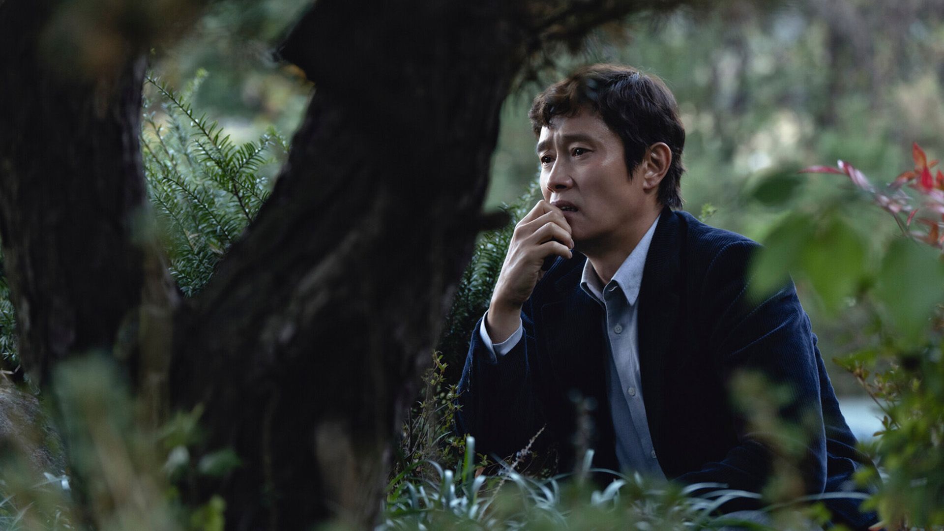 Lee Byung-hun w niebieskiej marynarce siedzi w trawie wśród drzew, na jego twarzy maluje się rozpacz