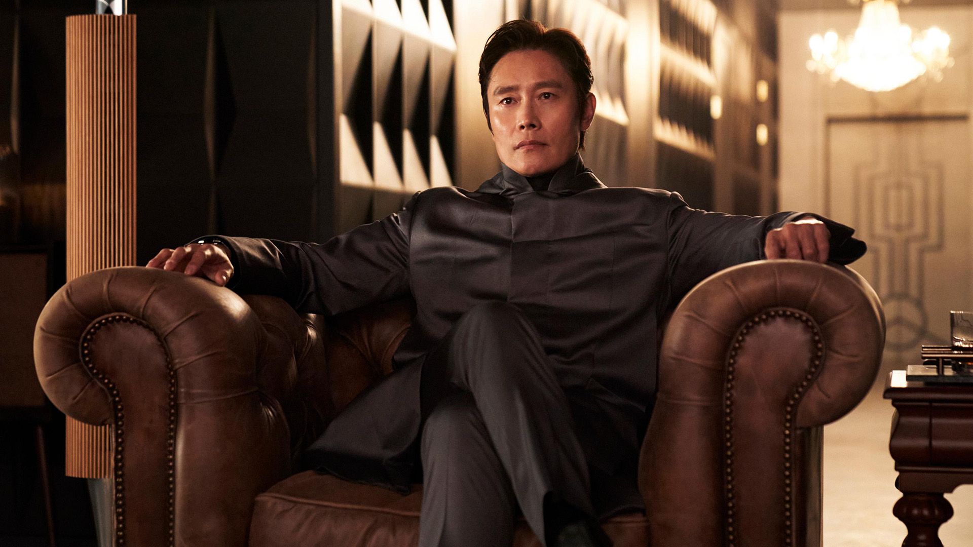Lee Byung-hun w szarym blezerze siedzi w skórzanym fotelu ze skrzyżowanymi nogami