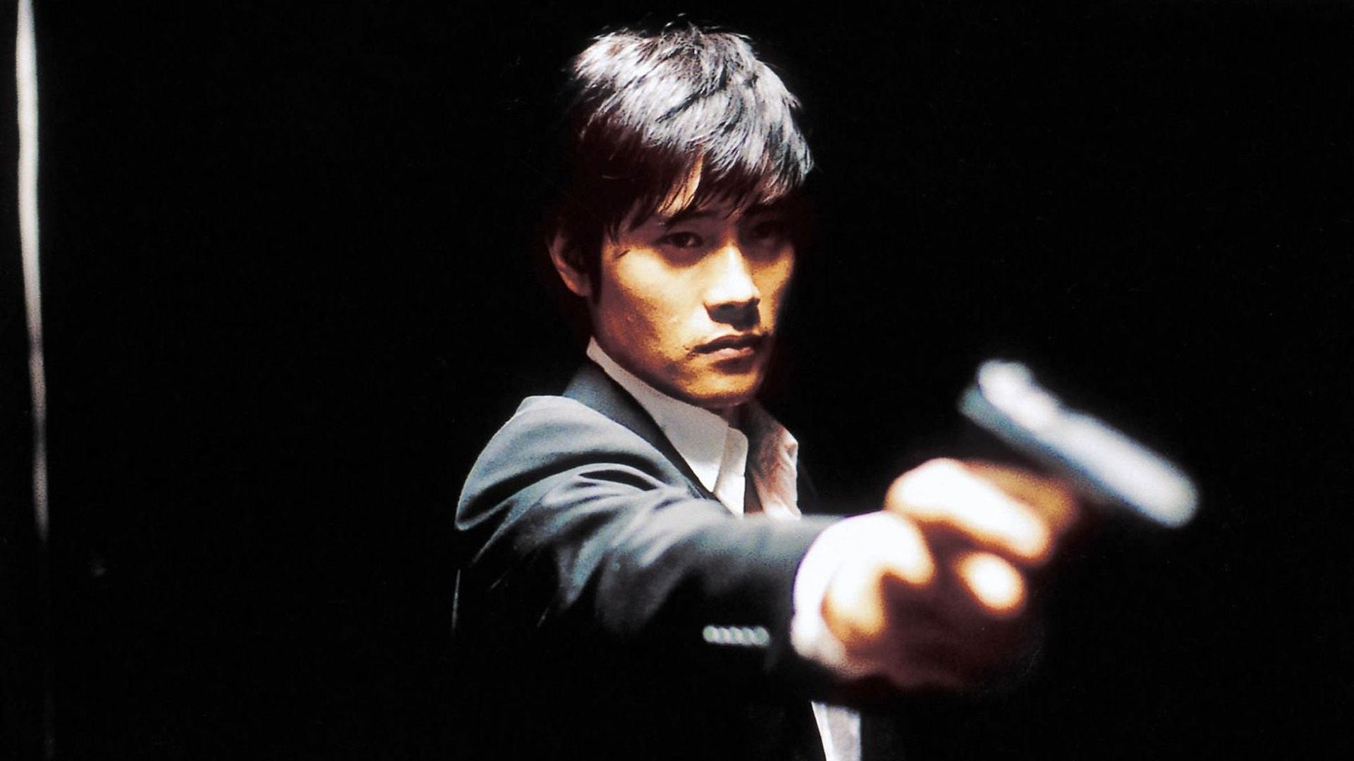 Lee Byung-hun w białej koszuli i marynarce trzyma pistolet w wyciągniętej dłoni