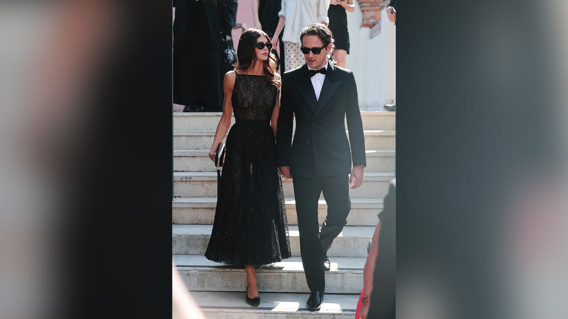 Lewis Pullman w czarnym smokingu i Kaia Gerber w czarnej wieczorowej sukni schodzą po schodach, oboje w czarnych okularach słonecznych