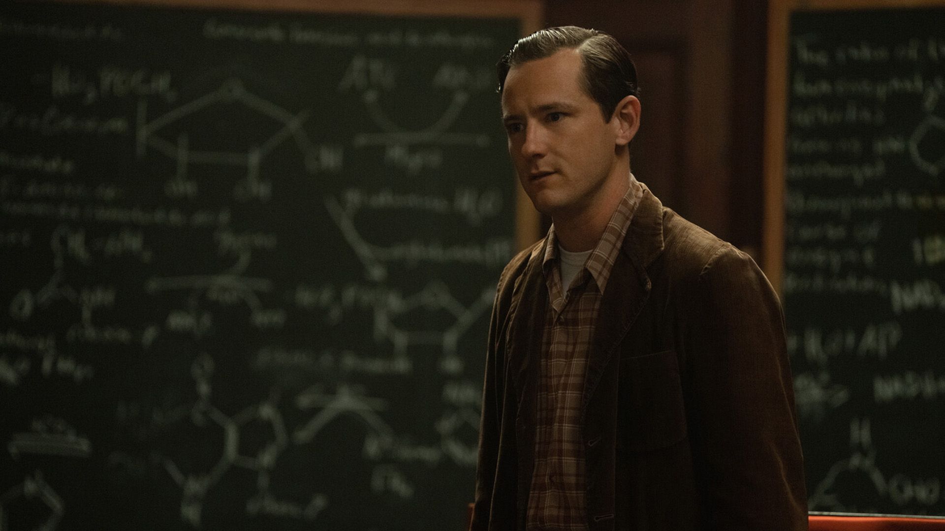 Lewis Pullman w brązowej sztruksowej marynarce i koszuli w kratkę stoi przy tablicy z wzorami chemicznymi