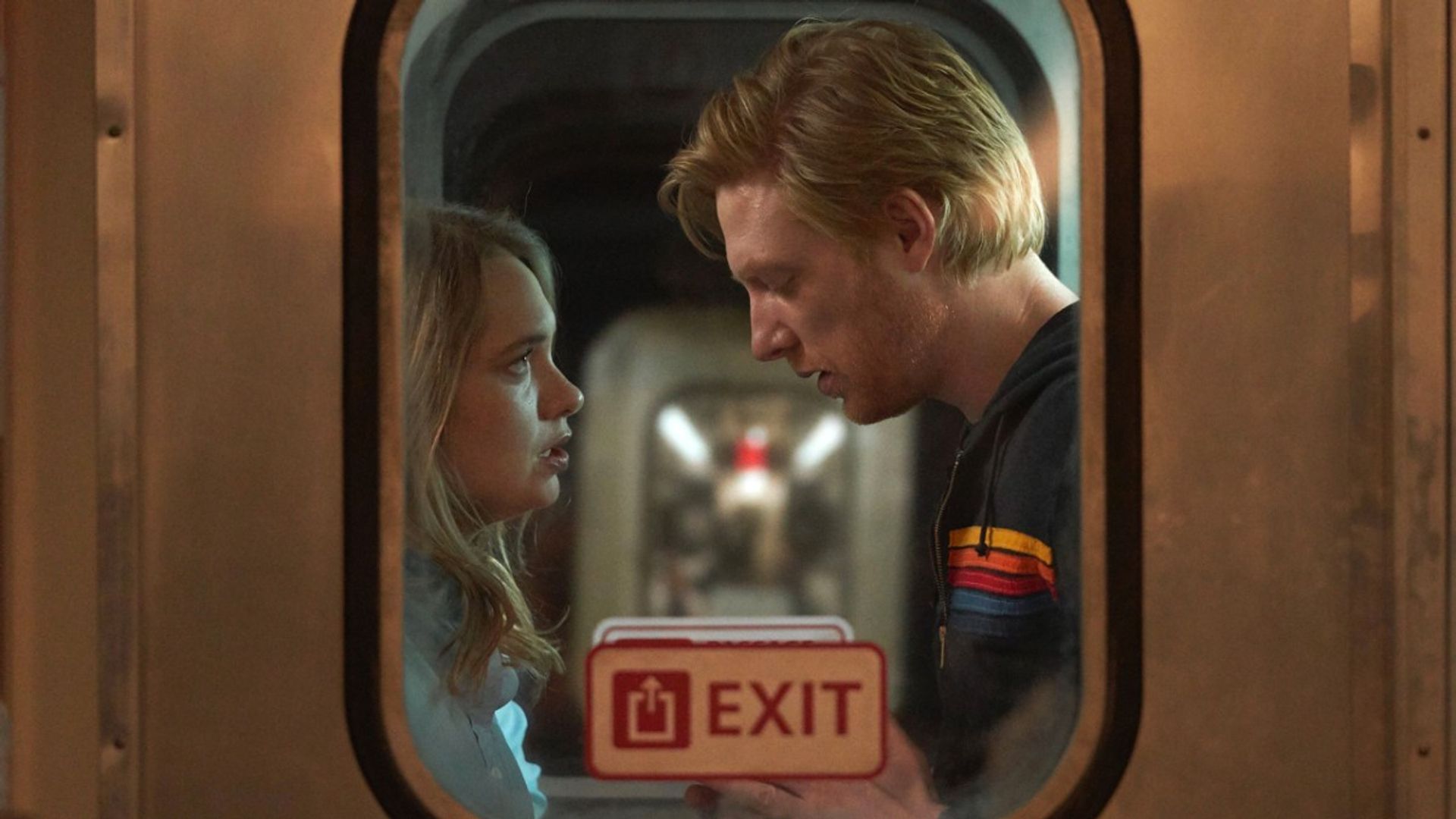 Domhnall Gleeson i Merritt Wever stoją twarzą w twarz w przedsionku pociągu, ujęcie z zewnątrz przez okno