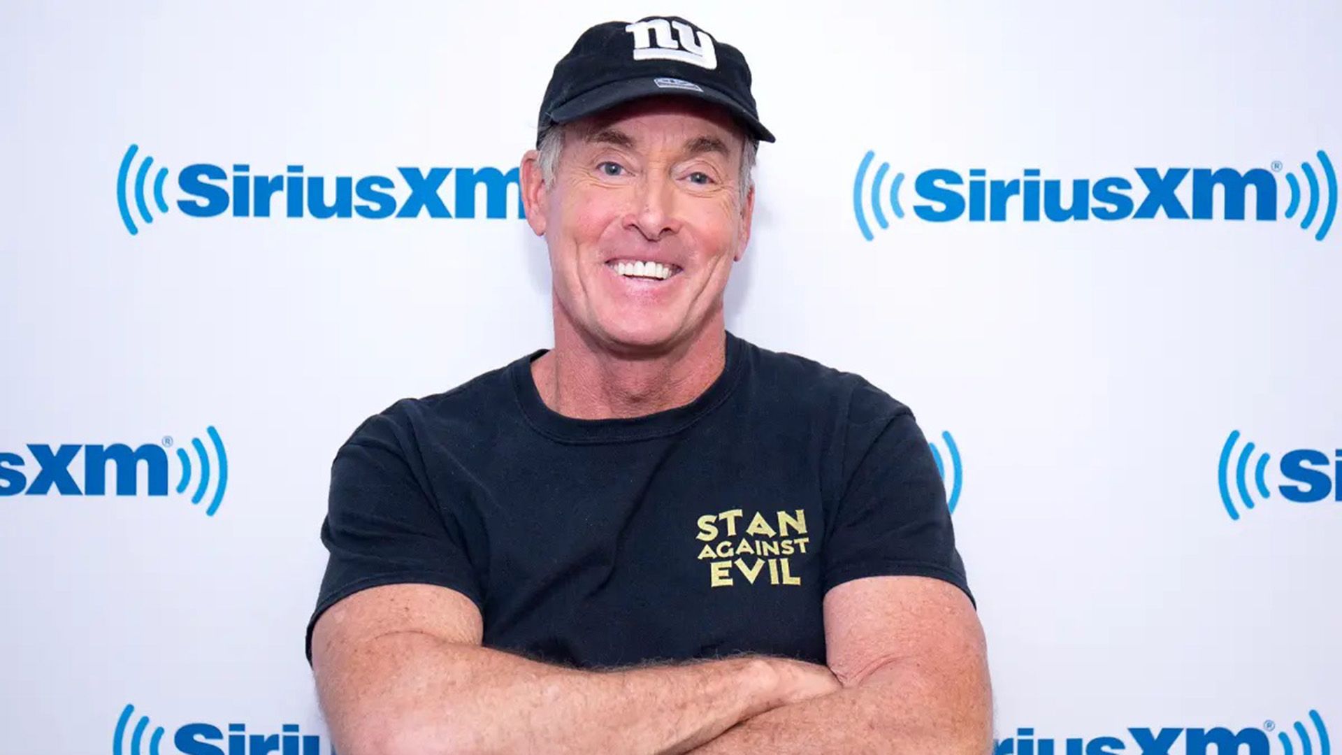 Aktor John C. McGinley w czapce z daszkiem, stojący ze skrzyżowanymi ramionami i szeroko się uśmiechający