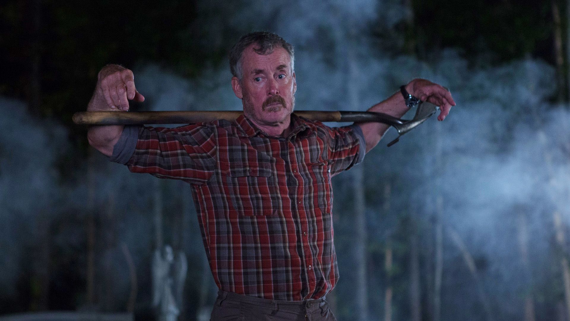 John C. McGinley w czerwonej koszuli w kratę stoi w przyćmionym świetle z łopatą na ramionach