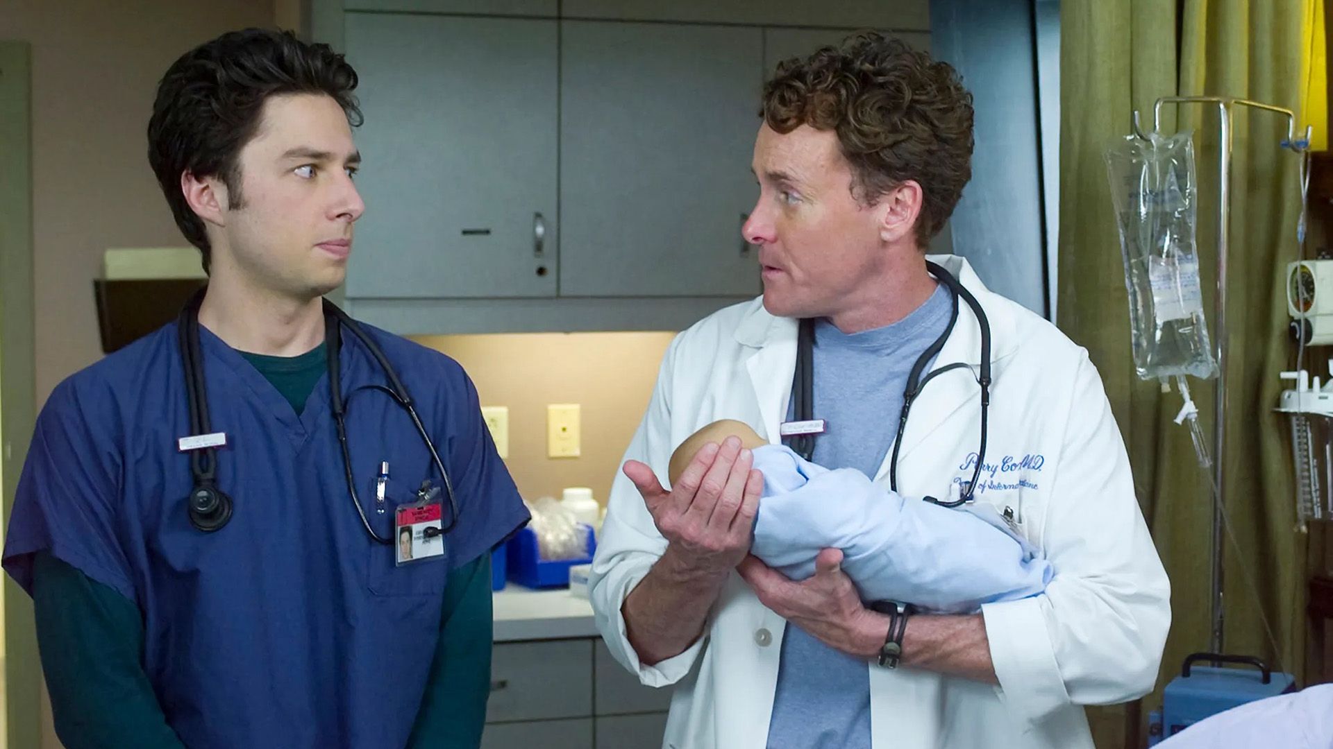 John C. McGinley w białym kitlu trzymający dziecko, obok stoi Zach Braff w niebieskim kitlu