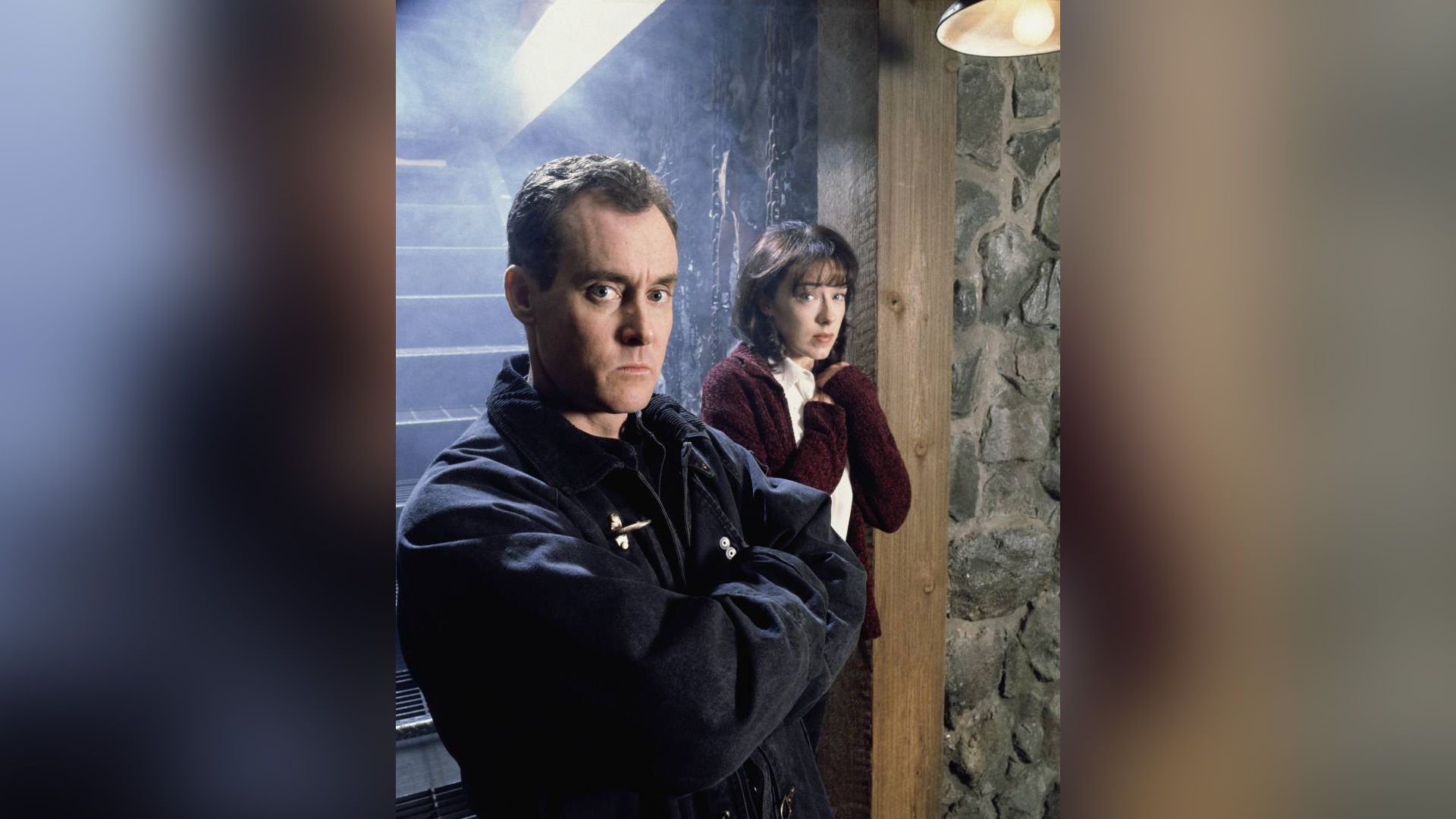 John C. McGinley z poważną miną stoi przy schodach, za nim przerażona dziewczyna