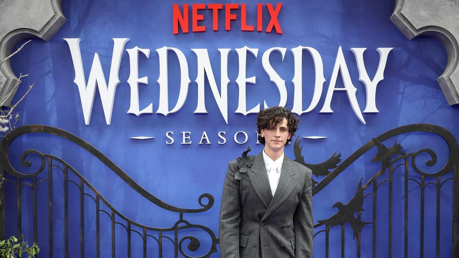 Owen Painter w garniturze stoi na tle ścianki Netflix z motywami Wednesday
